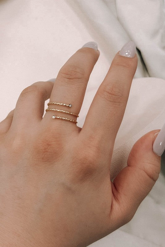 The Round and Round Ring boasts a stunning gold spiral design embellished with two small diamond accents, crafted from stainless steel with 14k gold plating, elegantly presented on a light beige background.