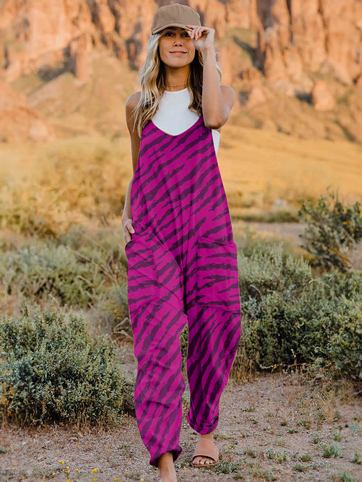 A person wearing a casual white V-neck tank top and a Full Size Printed V-Neck Sleeveless Jumpsuit with a brown animal print zebra-stripe pattern stands outdoors with one hand on their hat and the other in their pocket, set against a rocky landscape backdrop.