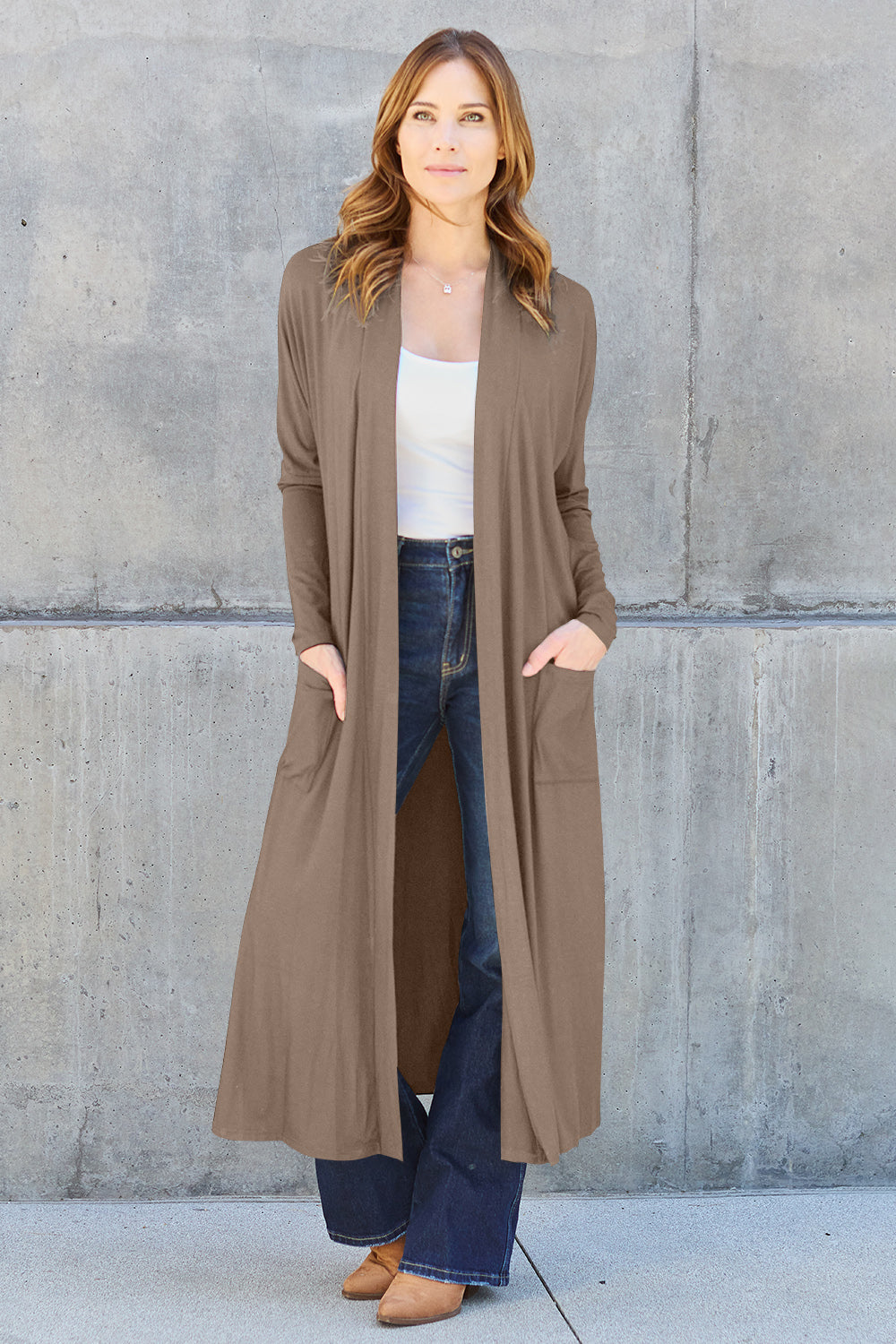 A woman stands against a concrete wall, wearing the Basic Bae Full Size Open Front Long Sleeve Cover Up made from rayon spandex, a white top, blue jeans in her perfect size, and brown shoes with her hands in her pockets. Machine wash cold for best results.