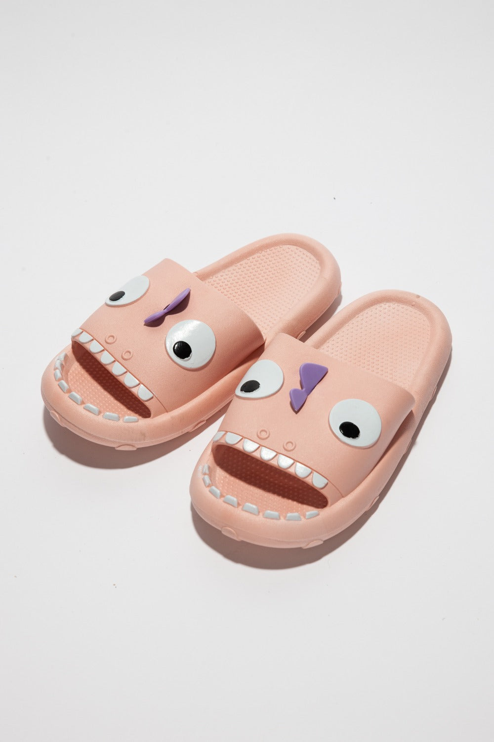 The NOOK JOI Monster Pillow Cloud Slides Non-Slip Slipper features cartoonish monster faces with large eyes, a purple nose, and an open mouth with teeth. Made of durable EVA material, these fun pink cloud slippers also boast an anti-slip design for added safety.