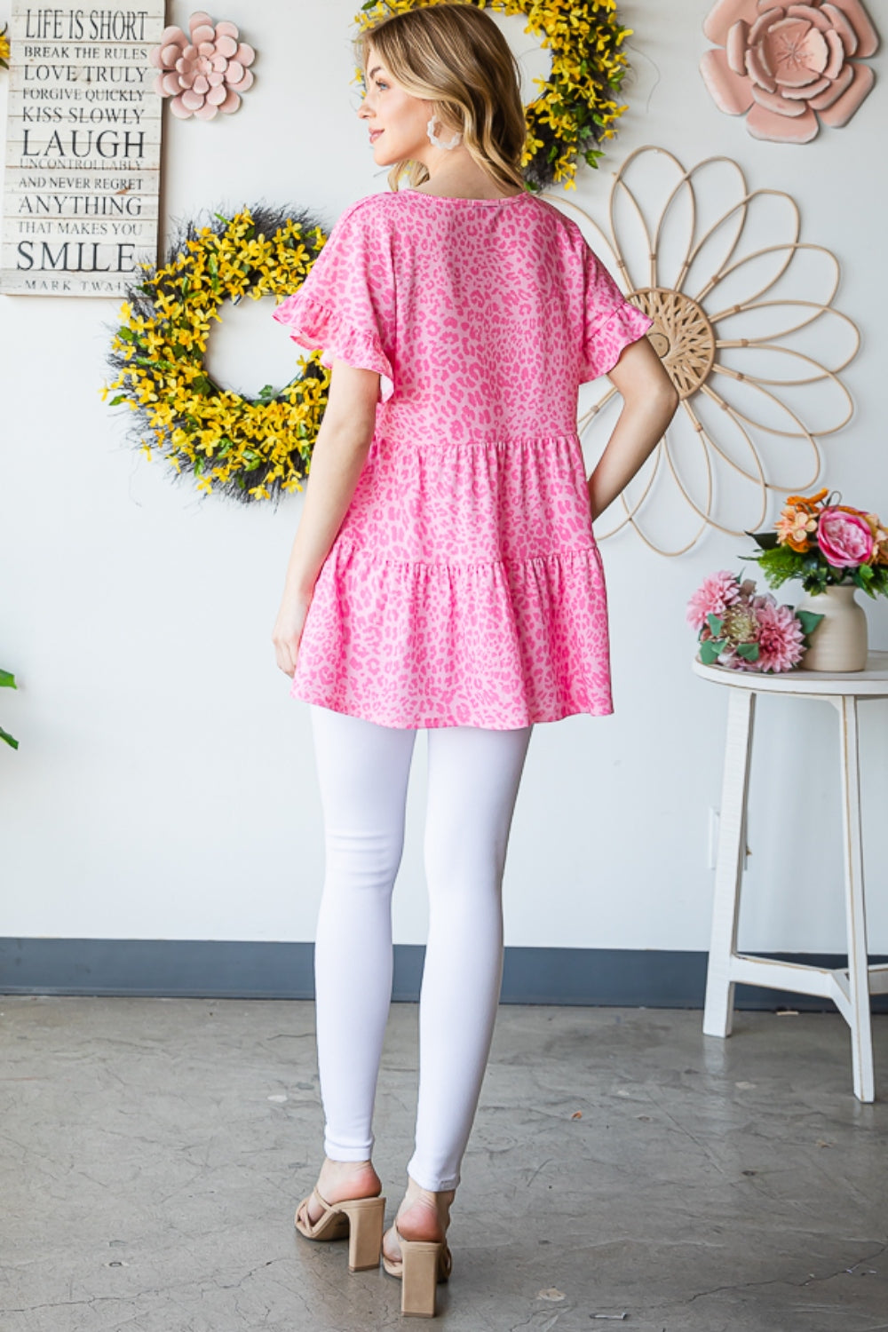 A person wearing a Heimish Full Size Leopard Ruffle Trim Short Sleeve Top stands in front of a floral wreath display.