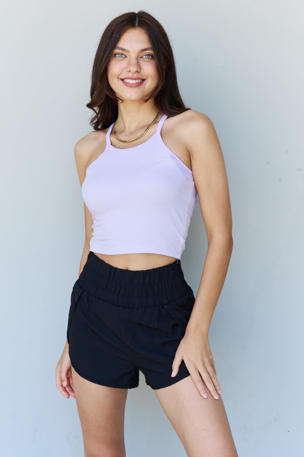 A woman with long brown hair, wearing the Ninexis Everyday Staple Soft Modal Short Strap Ribbed Tank Top in Lavender and black shorts, poses while smiling in front of a plain background.