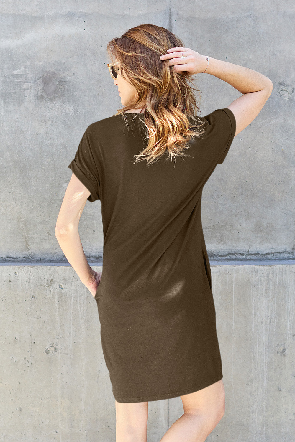 A woman wearing a dark olive-green Basic Bae Full Size Round Neck Short Sleeve Dress with Pockets and sunglasses stands against a concrete wall, holding a light-colored backpack.