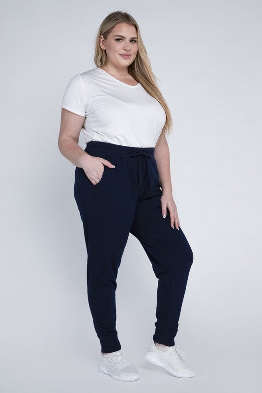 A person stands against a white background, wearing a white t-shirt, grey Plus-Size Jogger Pants with an adjustable waistband, and white sneakers. They have long blonde hair and are posing with one hand on their hip.