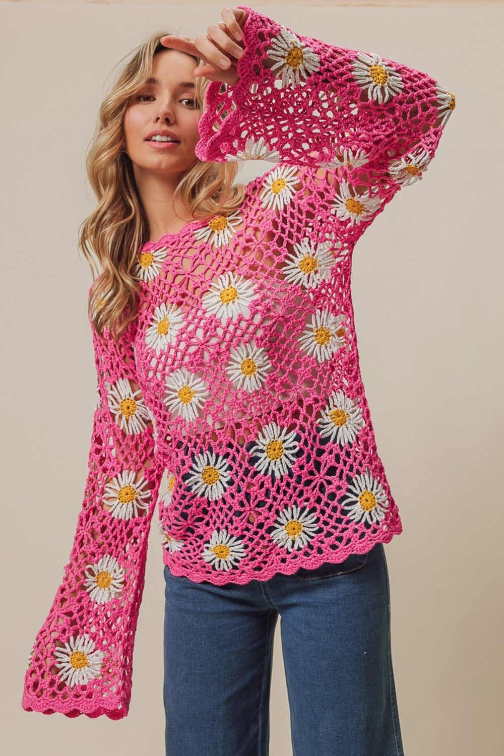 A person stands wearing the BiBi Floral Crochet Net Lace Cover Up, featuring pink crochet work adorned with white and yellow daisy patterns, exuding Bohemian style. They have long, wavy hair and pose with hands together.