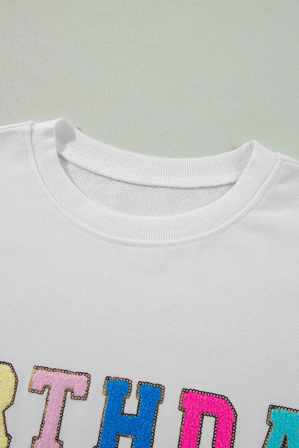 A person with long blonde hair is wearing a White BIRTHDAY QUEEN Graphic Balloon Sleeve Sweatshirt, paired with a pink feathered skirt, and sitting against a beautifully decorated backdrop.