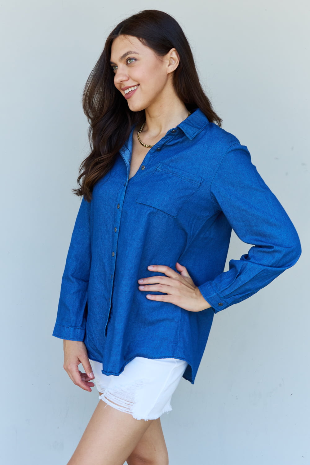 A person standing against a plain background, wearing the Doublju Blue Jean Baby Denim Button Down Shirt Top in Dark Blue and white shorts, smiling and holding a hand to their hair.