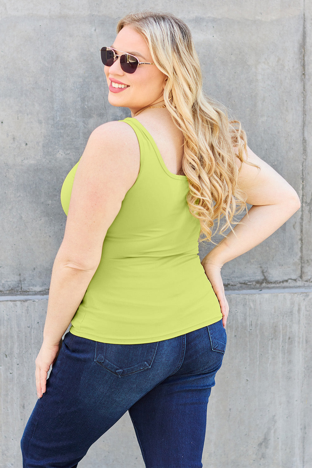 A woman with brown hair, wearing sunglasses, blue jeans, and the Basic Bae Full Size Square Neck Wide Strap Tank in black stands against a concrete wall, showcasing a basic style.