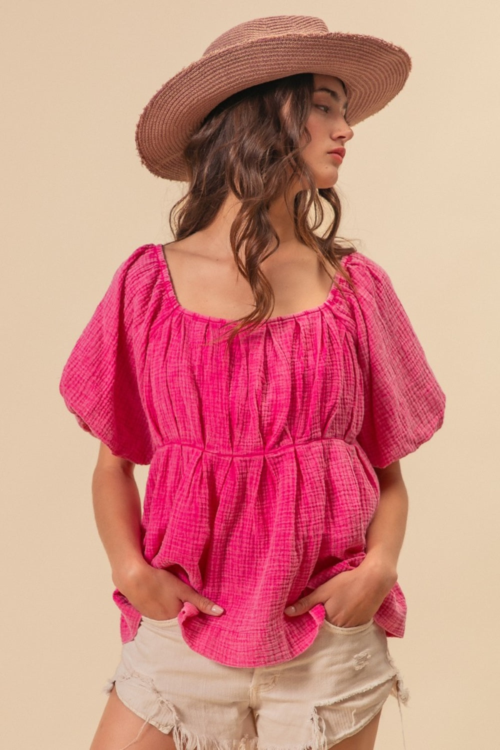 A woman wearing the BiBi Pleated Puff Sleeve Washed Blouse in pink, along with a wide-brimmed hat, strikes a pose against a beige background with one hand resting in her pocket.