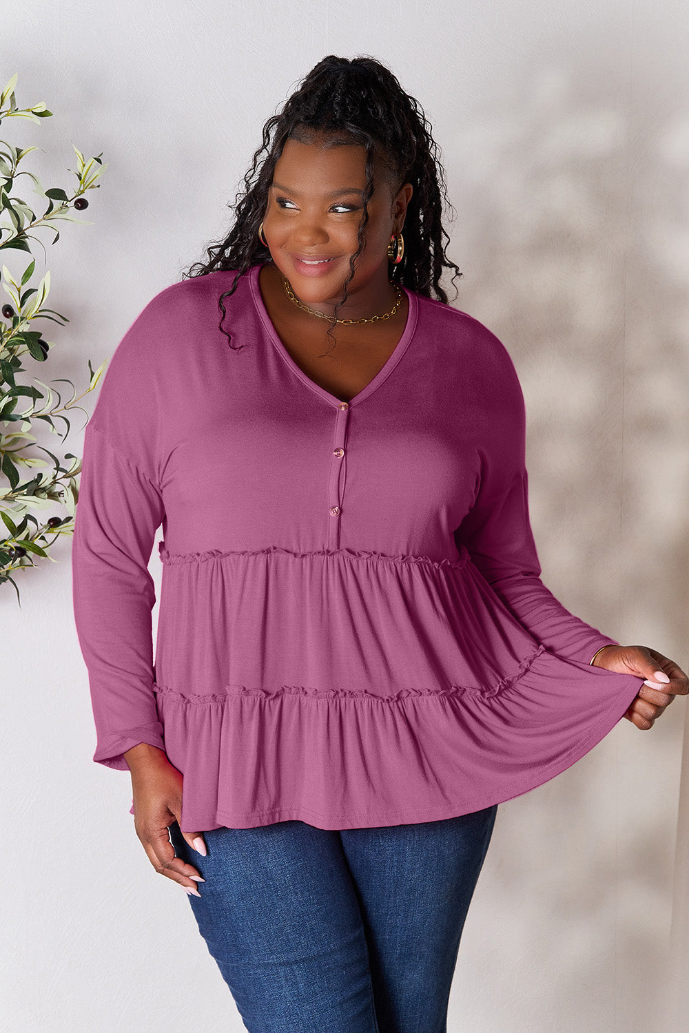 Person stands indoors near a plant, wearing the Double Take Half Button Long Sleeve Ruffle Hem Blouse in gray and blue jeans, smiling while looking to the side. The slightly stretchy fabric of the blouse adds comfort while its buttoned design offers a touch of elegance.