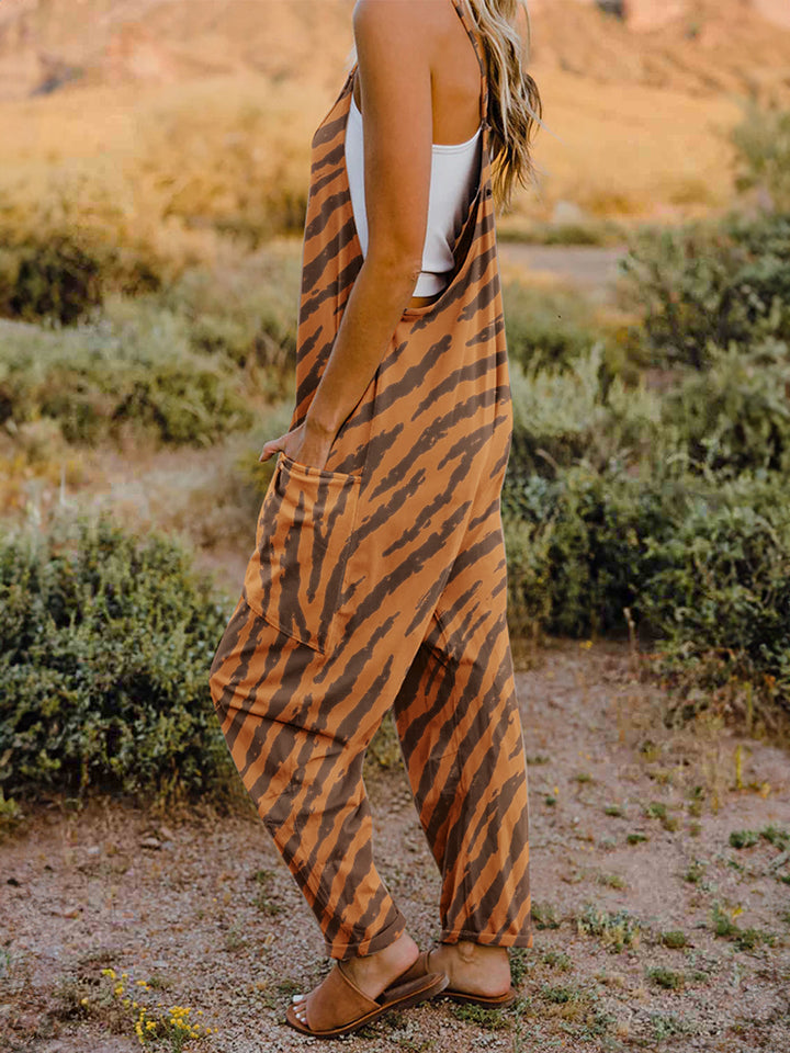 A person wearing a casual white V-neck tank top and a Full Size Printed V-Neck Sleeveless Jumpsuit with a brown animal print zebra-stripe pattern stands outdoors with one hand on their hat and the other in their pocket, set against a rocky landscape backdrop.