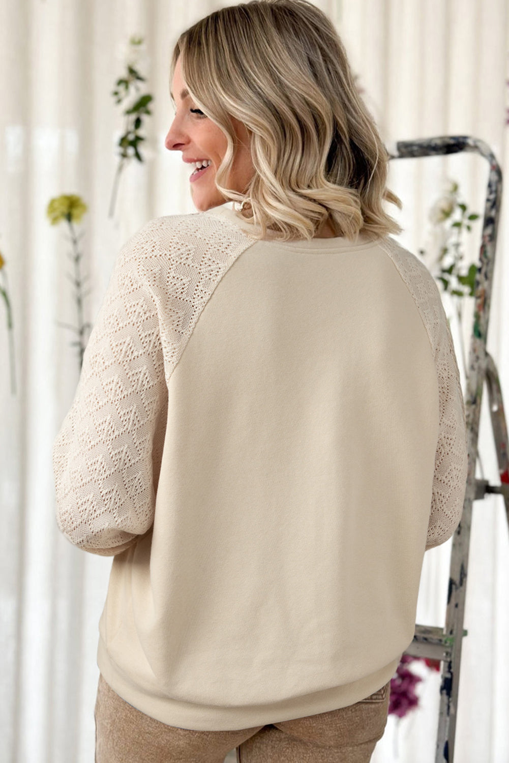 A blonde-haired individual wearing a Parchment Eyelet Knit Patchwork Raglan Sleeve Pullover Top stands indoors, smiling and looking to the side. Flowers and a ladder can be seen in the background.
