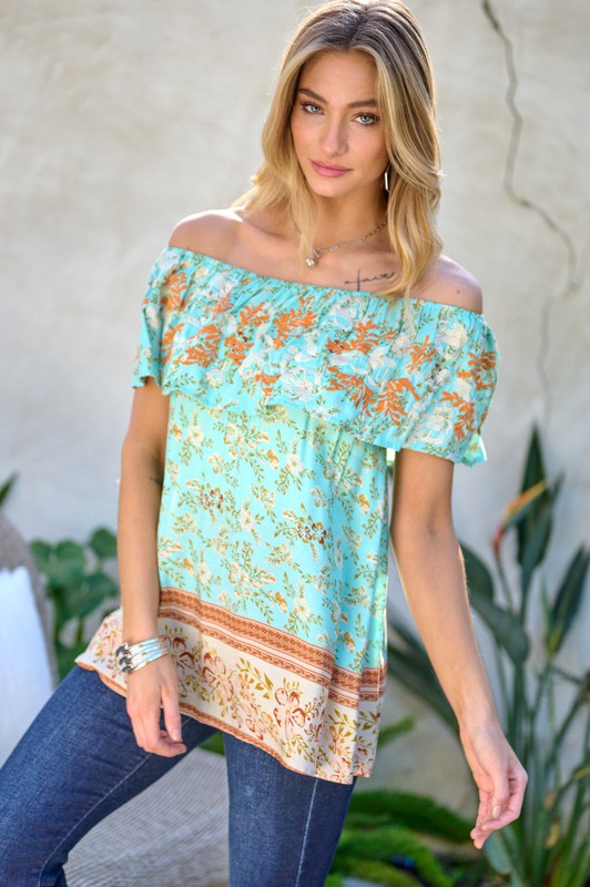 A woman with blonde hair is smiling while standing outdoors, wearing the Printed Off Shoulder Smocked Top paired with jeans.