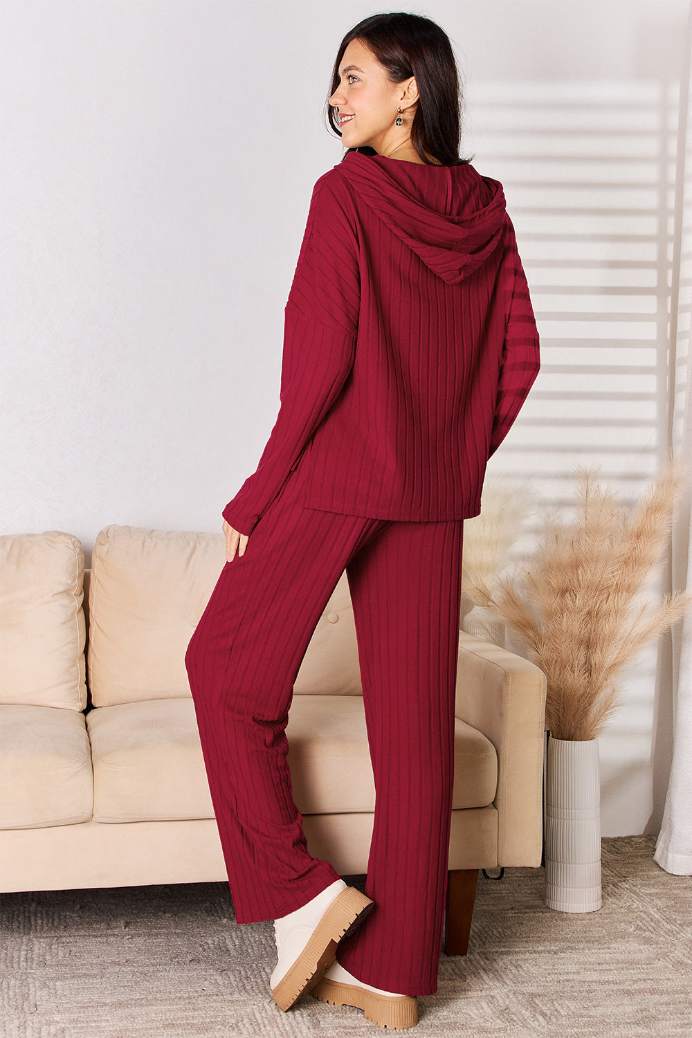 A person is standing indoors wearing a matching Basic Bae Full Size Ribbed Drawstring Hooded Top and Straight Pants Set in maroon, paired with white shoes. Behind them, there is a cream-colored sofa and a vase with beige decorative grass. The outfit, perfect for any casual day, boasts an imported quality and is best maintained with a machine wash cold care routine.