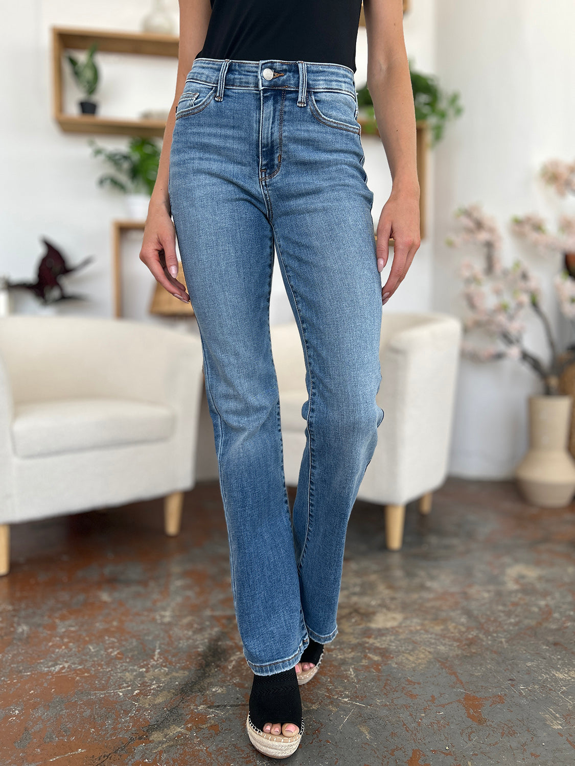 Person wearing Judy Blue Full Size Mid-Rise Waist Straight Jeans and a black top, standing in a room with white chairs, potted plants, and wooden shelves. Only the lower half of the body and part of the arms are visible.