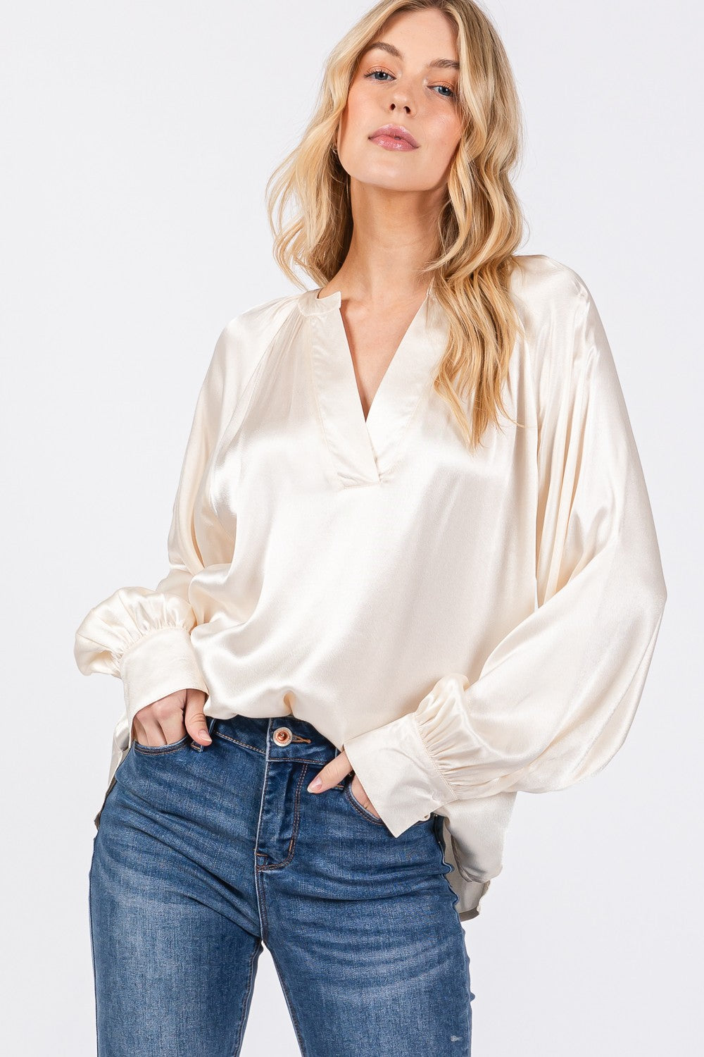 A person is wearing a SAGE + FIG Notched Long Sleeve Blouse in cream-colored satin paired with blue jeans.