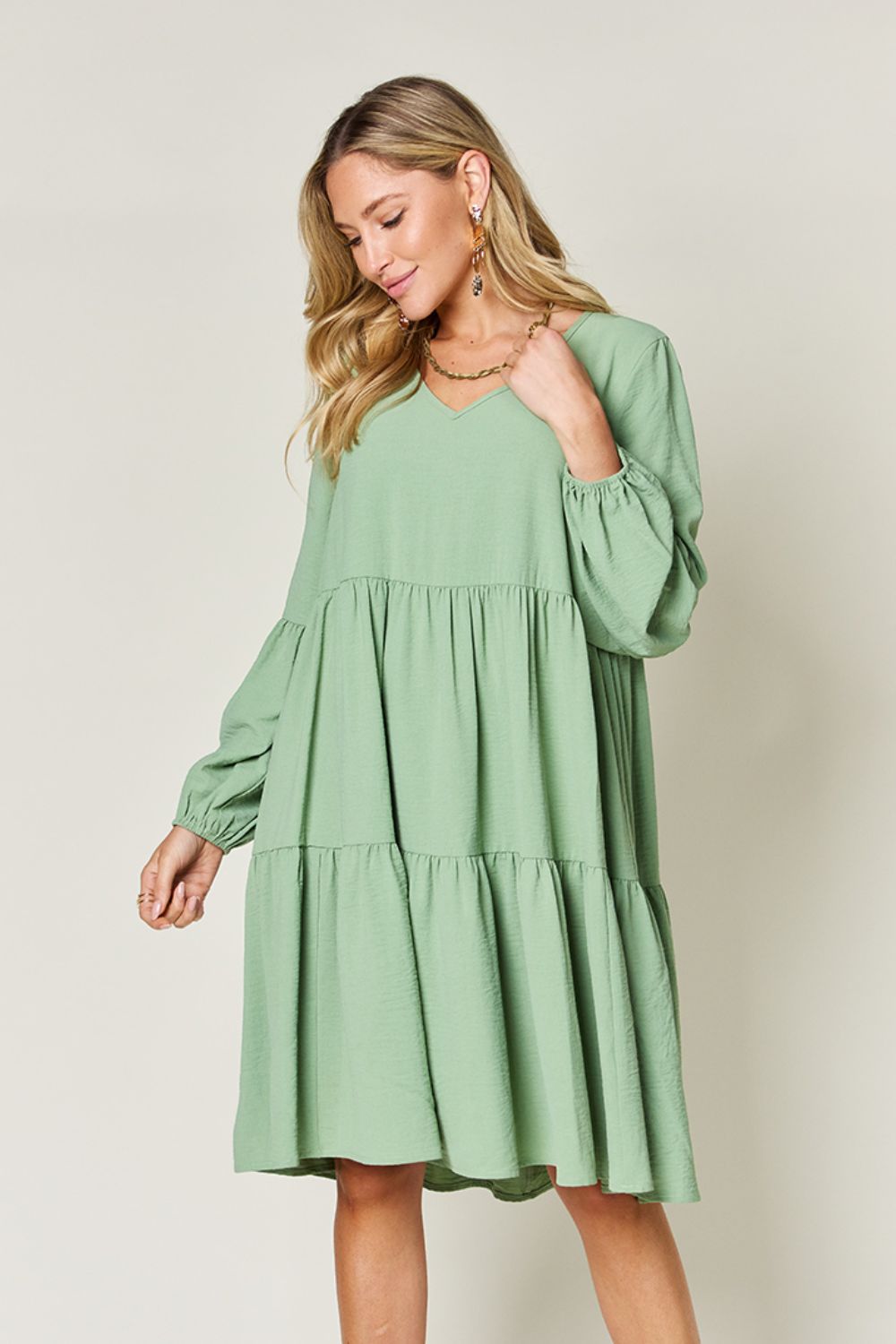 A woman stands wearing the Double Take Full Size V-Neck Balloon Sleeve Tiered Dress with Pockets in pink. Crafted from high stretch yarn, this dress combines comfort and style effortlessly. She has long, wavy hair and accessorizes with hoop earrings and a necklace. This imported garment is easy to care for – simply machine wash cold to keep it looking fresh.