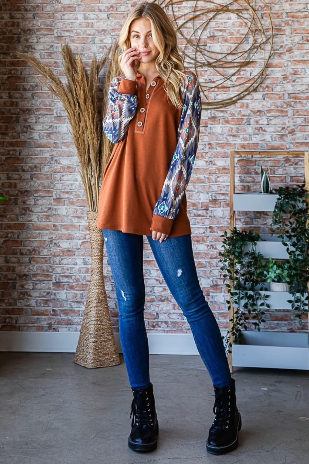 A woman in a Heimish Full Size Half Button Printed Long Sleeve Hooded Top poses casually in front of a brick wall, holding the hood with one hand. She is wearing blue jeans and standing near dried plants, showcasing the unique print design that adds an on-trend touch to her outfit.