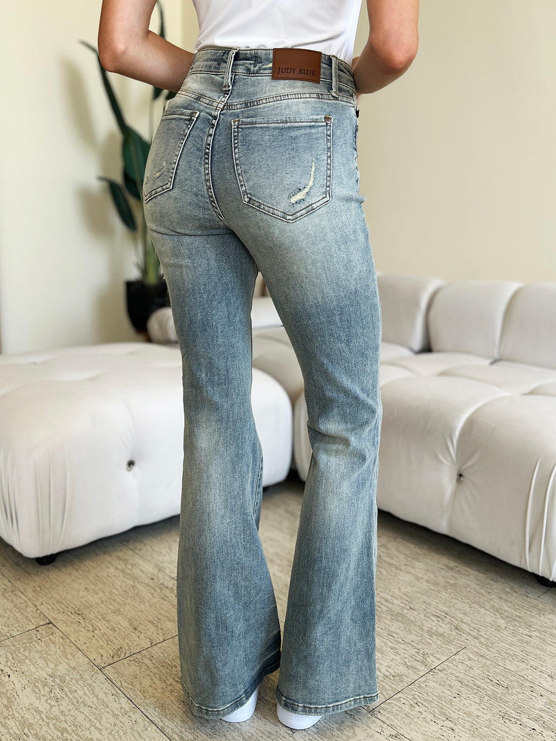 A person wearing Judy Blue Full Size Mid Rise Flare Jeans in a retro-inspired light blue color, paired with a white top and white shoes, stands in a modern living room with a white tufted sofa in the background.