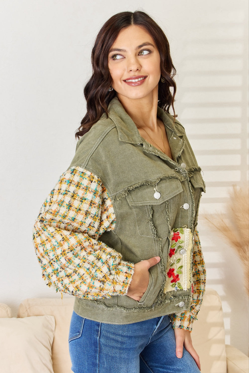 A woman with long hair stands indoors, wearing the POL Embroidered Button Down Raw Hem Shacket with floral designs and contrast tweed sleeves, paired with blue jeans. She has one hand touching her hair and the other resting in her jacket pocket.