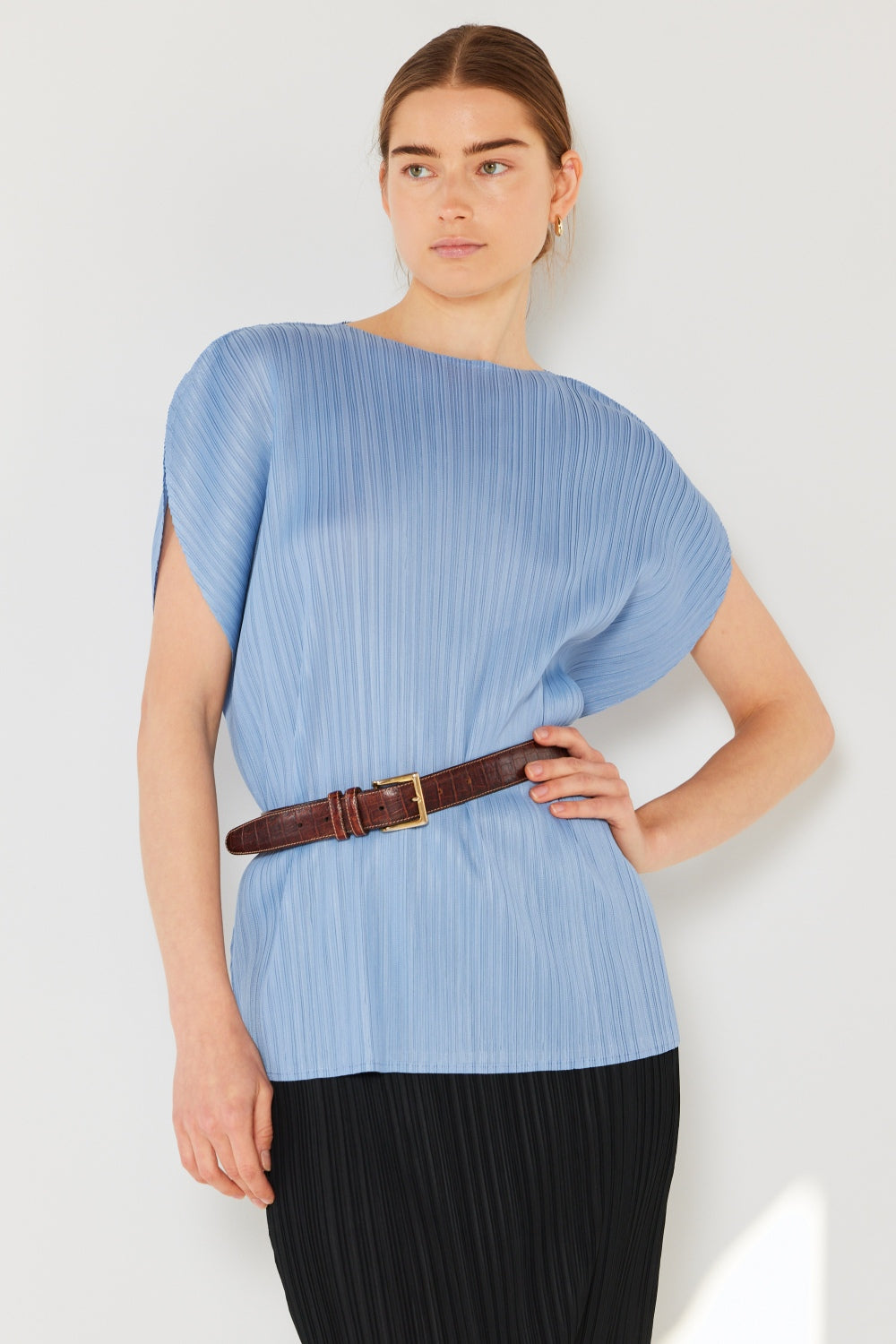 A person wearing the Marina West Swim Rib Pleated Oversized Dolman Sleeve Top in a fashionable light blue shade, paired with a brown belt and black skirt, stands against a plain background.