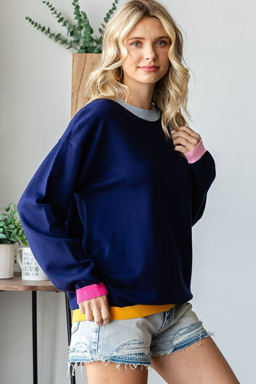 A person with long blonde hair wearing a First Love Contrast Ribbed Round Neck Long Sleeve Sweater and light denim shorts stands indoors near a small table and plant.