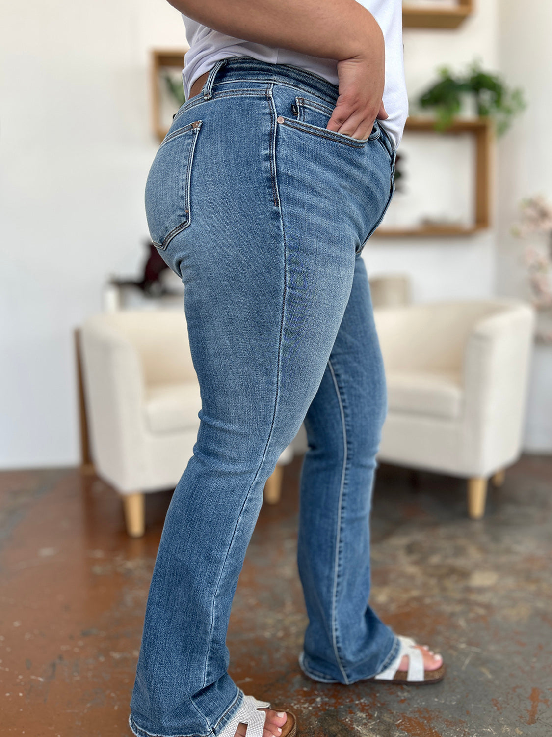 Person wearing Judy Blue Full Size Mid-Rise Waist Straight Jeans and a black top, standing in a room with white chairs, potted plants, and wooden shelves. Only the lower half of the body and part of the arms are visible.