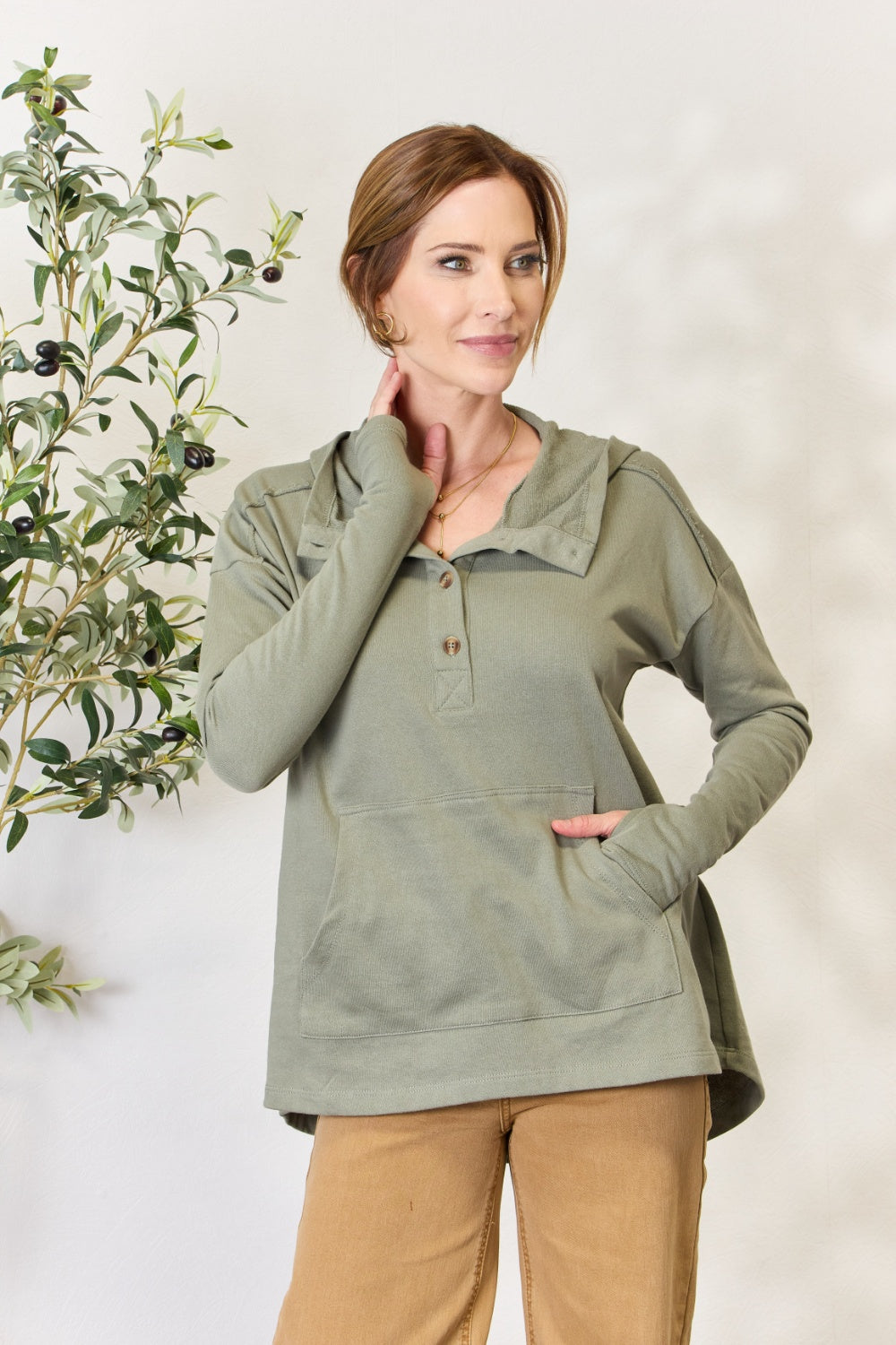 A person stands with hands in the pockets of their light green Culture Code Full Size Half Button Hoodie and brown pants. A plant is visible on the left side of the image.