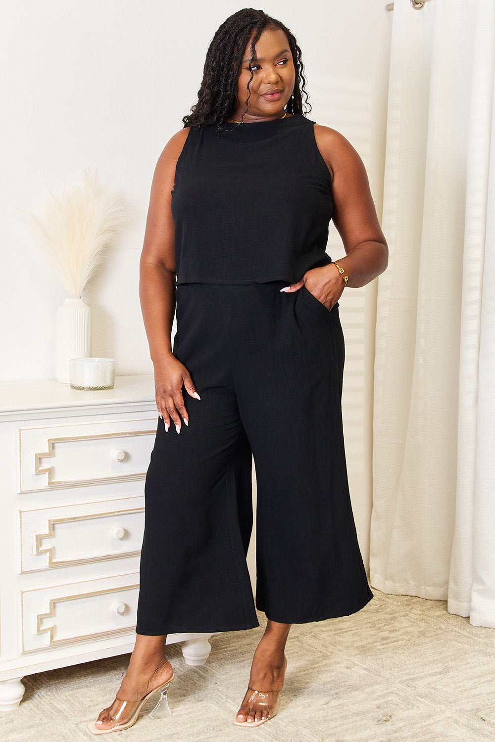 A woman in a stylish Devine Buttoned Round Neck Tank and Wide Leg Pants Set, featuring hands in pockets, smiles near a white dresser adorned with decorative items.