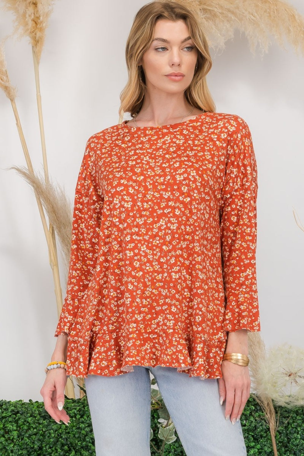 A woman wearing the Celeste Full Size Floral Ruffle Detail Top and jeans stands smiling in front of pampas grass and greenery, exuding feminine flair.