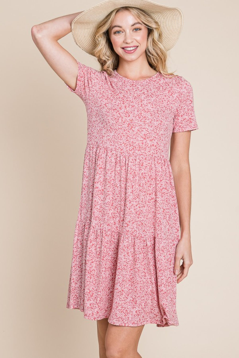 A woman with wavy blonde hair is smiling and holding the side of her BOMBOM Printed Short Sleeve Mini Dress, a lightweight summer dress featuring a pink pattern.