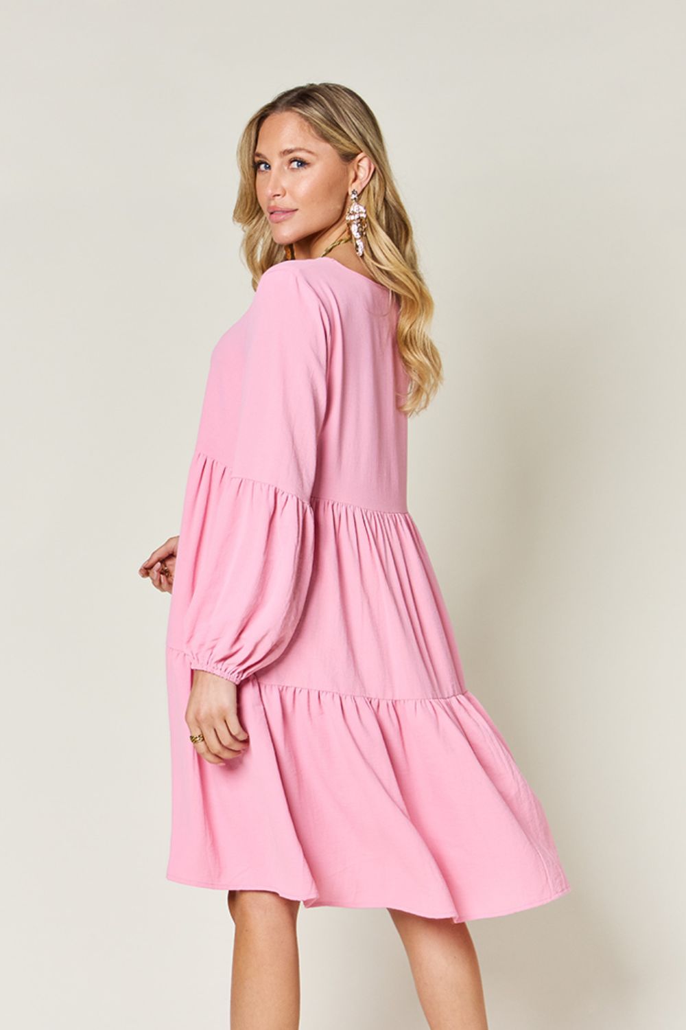 A woman stands wearing the Double Take Full Size V-Neck Balloon Sleeve Tiered Dress with Pockets in pink. Crafted from high stretch yarn, this dress combines comfort and style effortlessly. She has long, wavy hair and accessorizes with hoop earrings and a necklace. This imported garment is easy to care for – simply machine wash cold to keep it looking fresh.