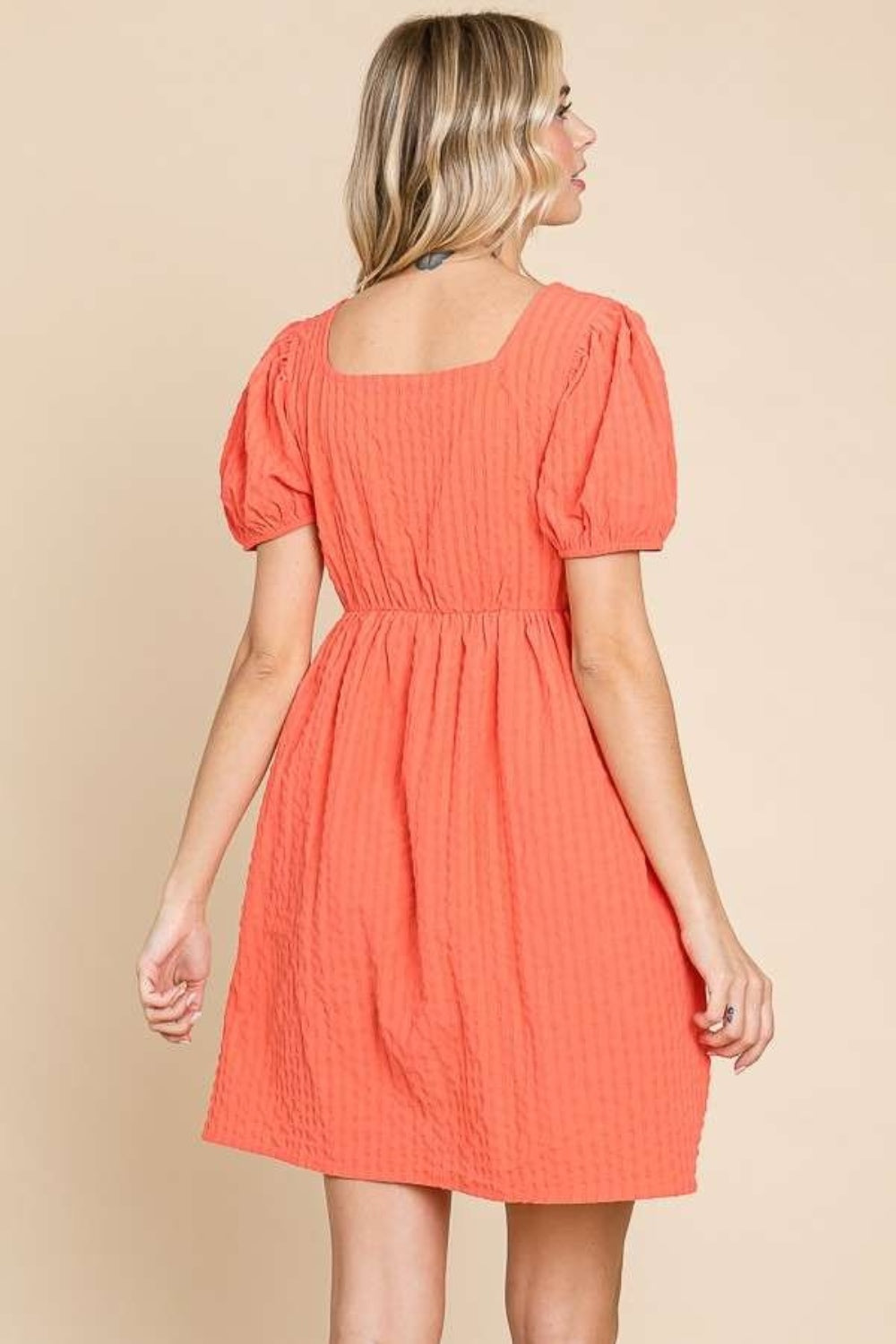 A woman is wearing a Culture Code Textured Square Neck Short Sleeve Dress: it's a stylish, short-sleeved, square-necked, orange dress with puffed sleeves. She is smiling and has one hand in her pocket.