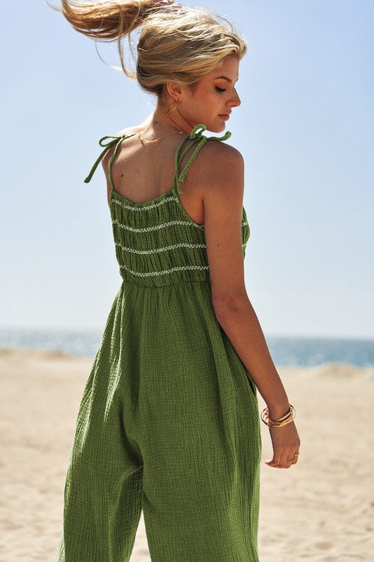 A person wearing the Washed Multi Smocked Detail Tie Straps Jumpsuit stands on the sunny beach, with the ocean creating a perfect backdrop.