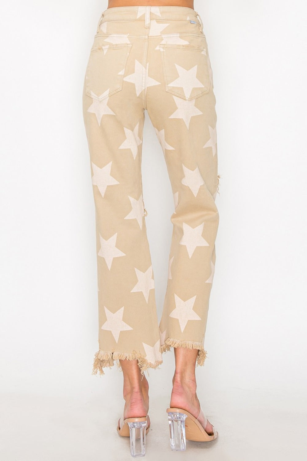 Someone dressed in RISEN Full Size High Rise Star Printed Frayed Hem Jeans with ripped knees, complemented by clear heels.