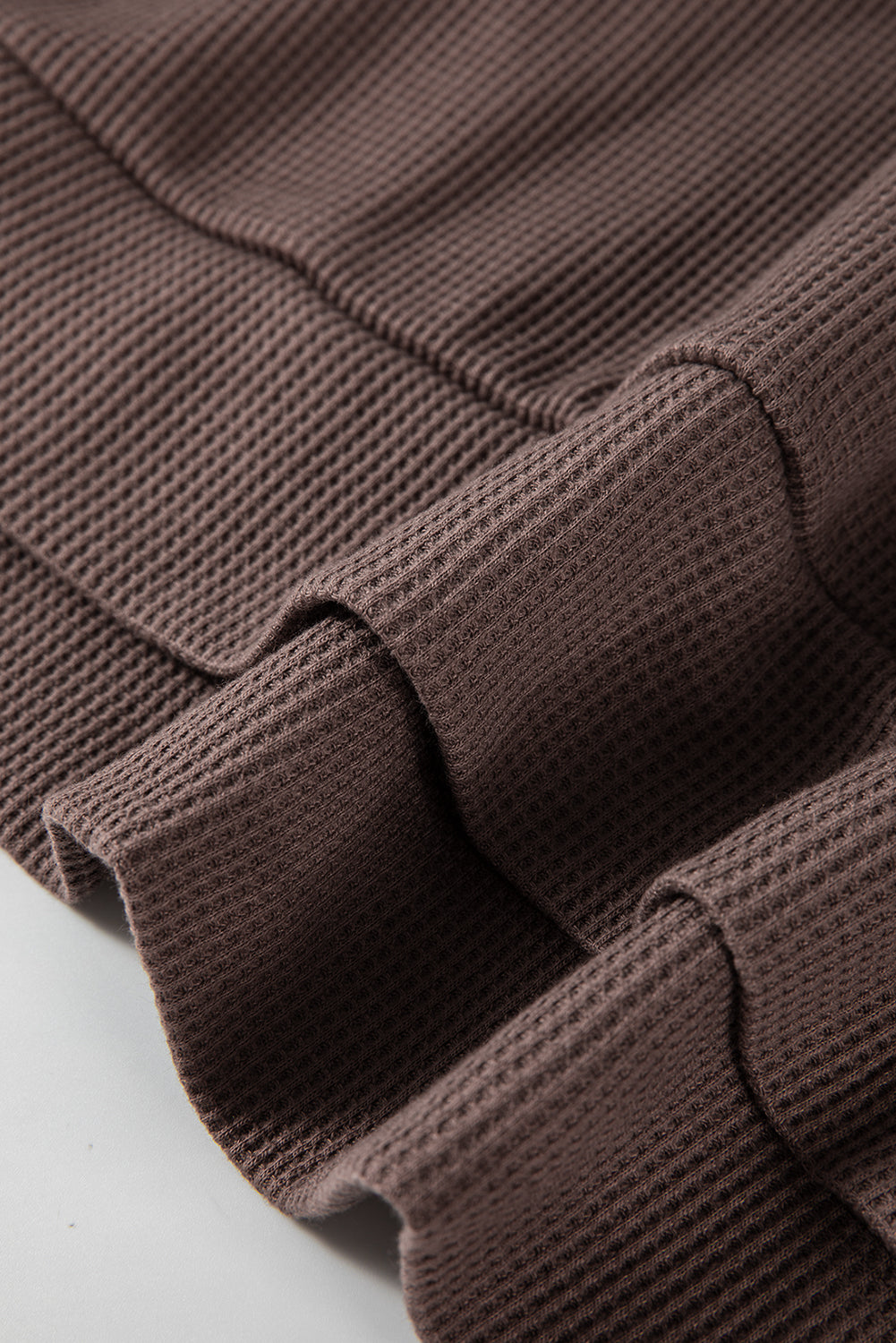 A woman with long hair is standing in profile, wearing a Coffee Waffle Knit Fleece Lined High Low Oversized Hoodie and black shorts. The background is a plain light gray.