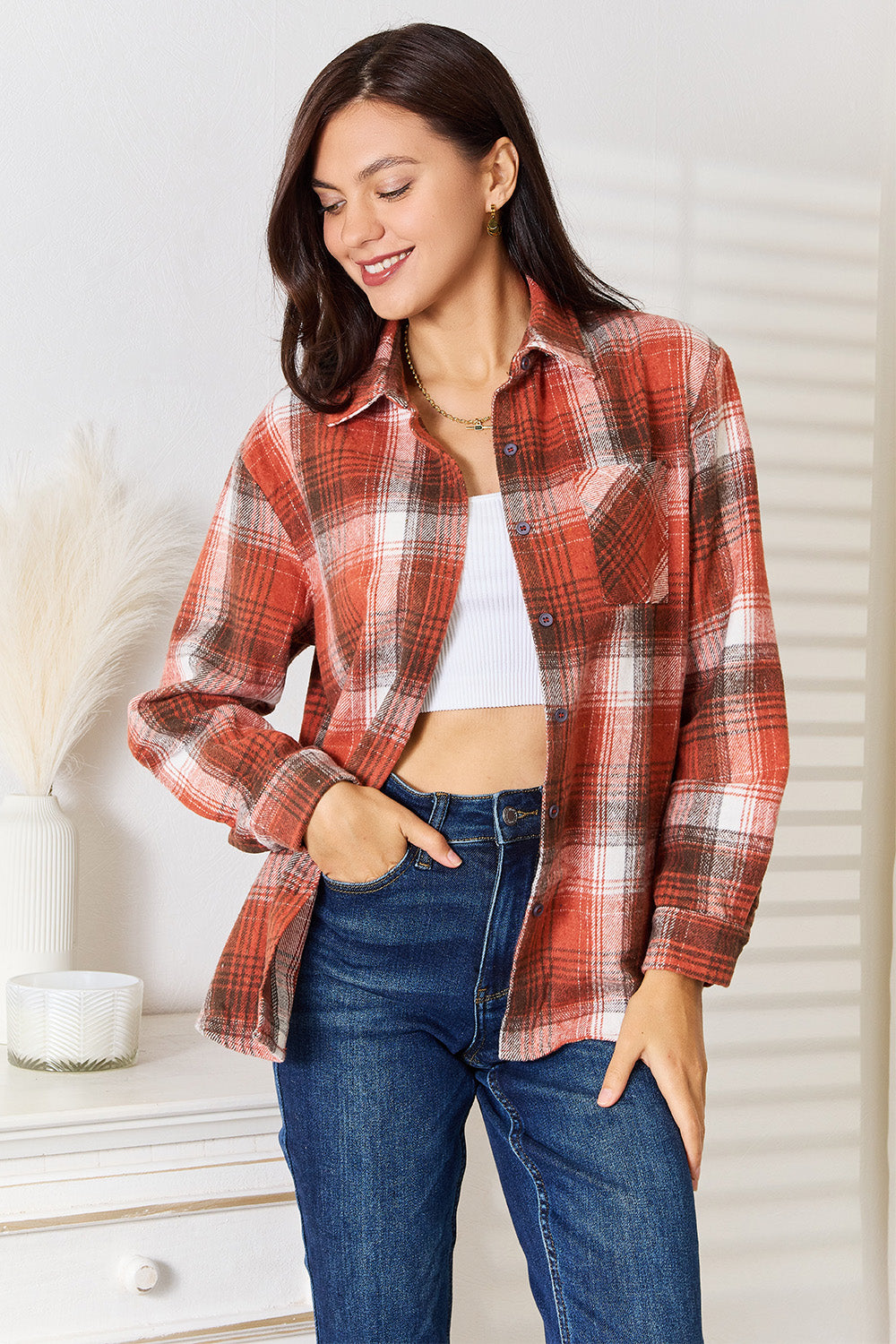 A woman in a Plaid Collared Neck Long Sleeve Shirt and blue jeans stands indoors, smiling and looking downward. She is wearing a white top underneath the classic wardrobe staple.