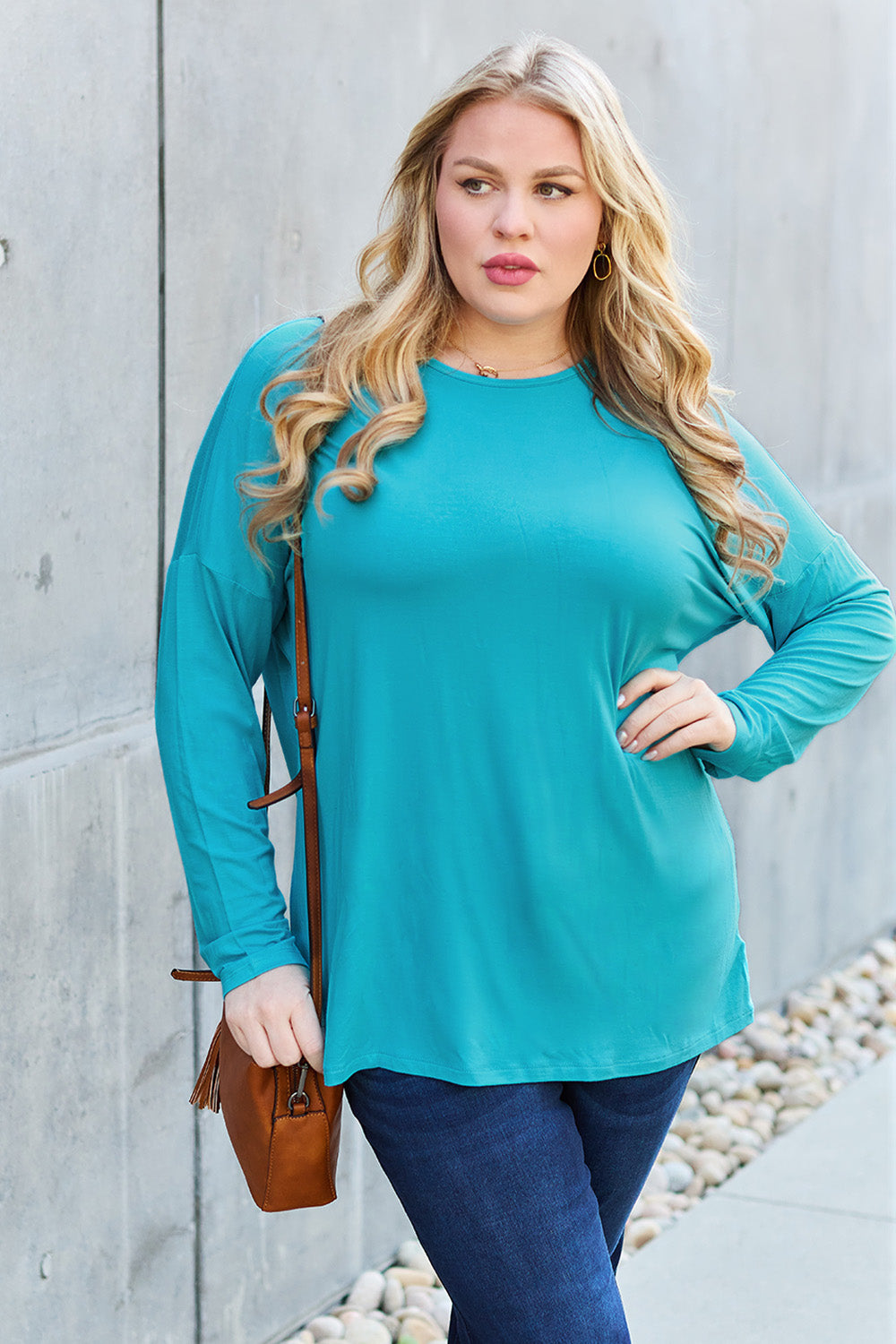 A woman with shoulder-length hair wearing a Basic Bae Full Size Round Neck Dropped Shoulder T-Shirt in green and a pair of jeans, exuding a basic style, stands against a concrete wall. She's holding an imported brown woven backpack on her shoulder while touching her hair with her hand.