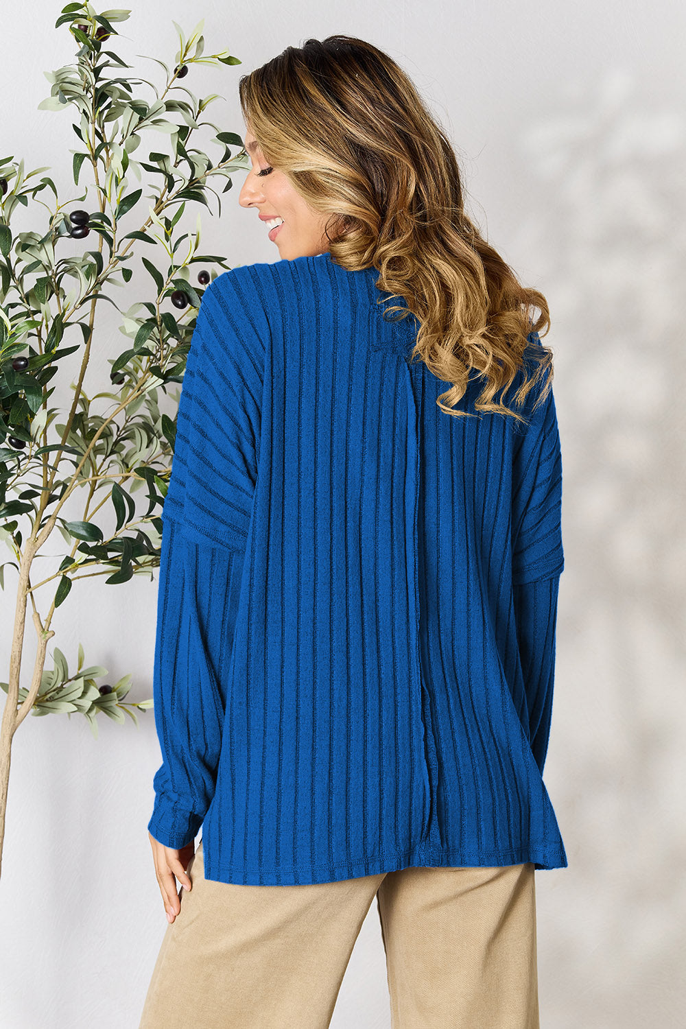 A person wearing the Basic Bae Full Size Ribbed Round Neck Slit Knit Top in blue and beige pants stands and smiles in front of a leafy plant.