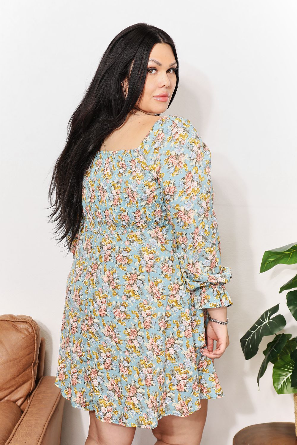 A woman in the Honey Floral Smocked Flounce Sleeve Square Neck Dress stands indoors, holding the hem of her dress. She is smiling slightly with her eyes looking downward. Nearby, a plant and a brown couch complete the cozy scene.