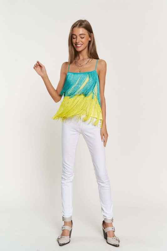 A woman with long brown hair is wearing a Fringe Overlay Cross Straps Party Cami Top featuring blue and yellow layers, paired with white pants, standing against a plain background.