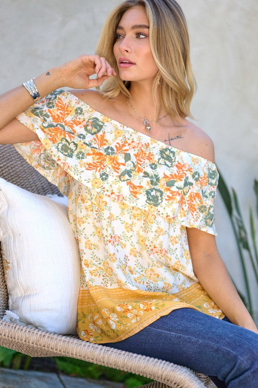 A woman with blonde hair is smiling while standing outdoors, wearing the Printed Off Shoulder Smocked Top paired with jeans.