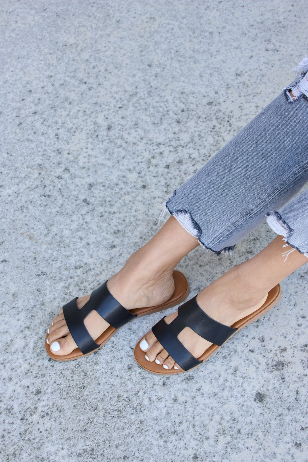 Someone wearing Forever Link Cutout Open Toe Flat Sandals and distressed jeans on a textured concrete surface.