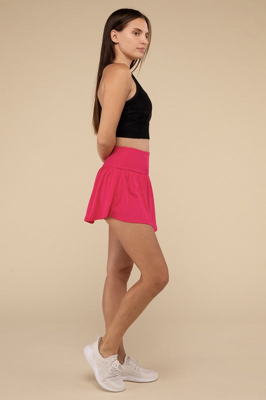 Close-up of a woman in a black crop top and Wide Band Tennis Skirt with Zippered Back Pocket, pulling the skirt to show matching shorts with a zippered back pocket underneath.