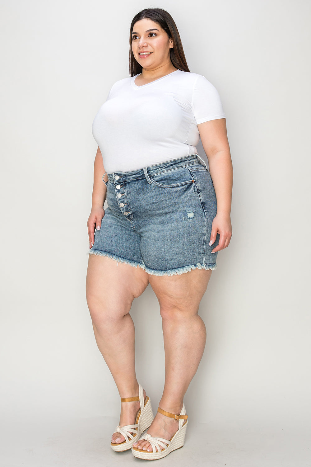 Wearing the Judy Blue Full Size Button Fly Raw Hem Denim Shorts and a white t-shirt, a person stands smiling in sandals against a plain background.