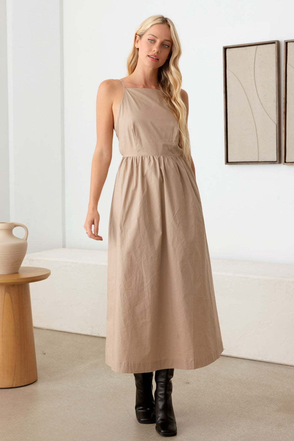 A woman with long blonde hair wears a beige Le Lis Tie Back Backless Cami Dress made from 100% cotton and black knee-high boots. She stands indoors next to a wooden stool with a ceramic vase, while simple wall art adorns the background.