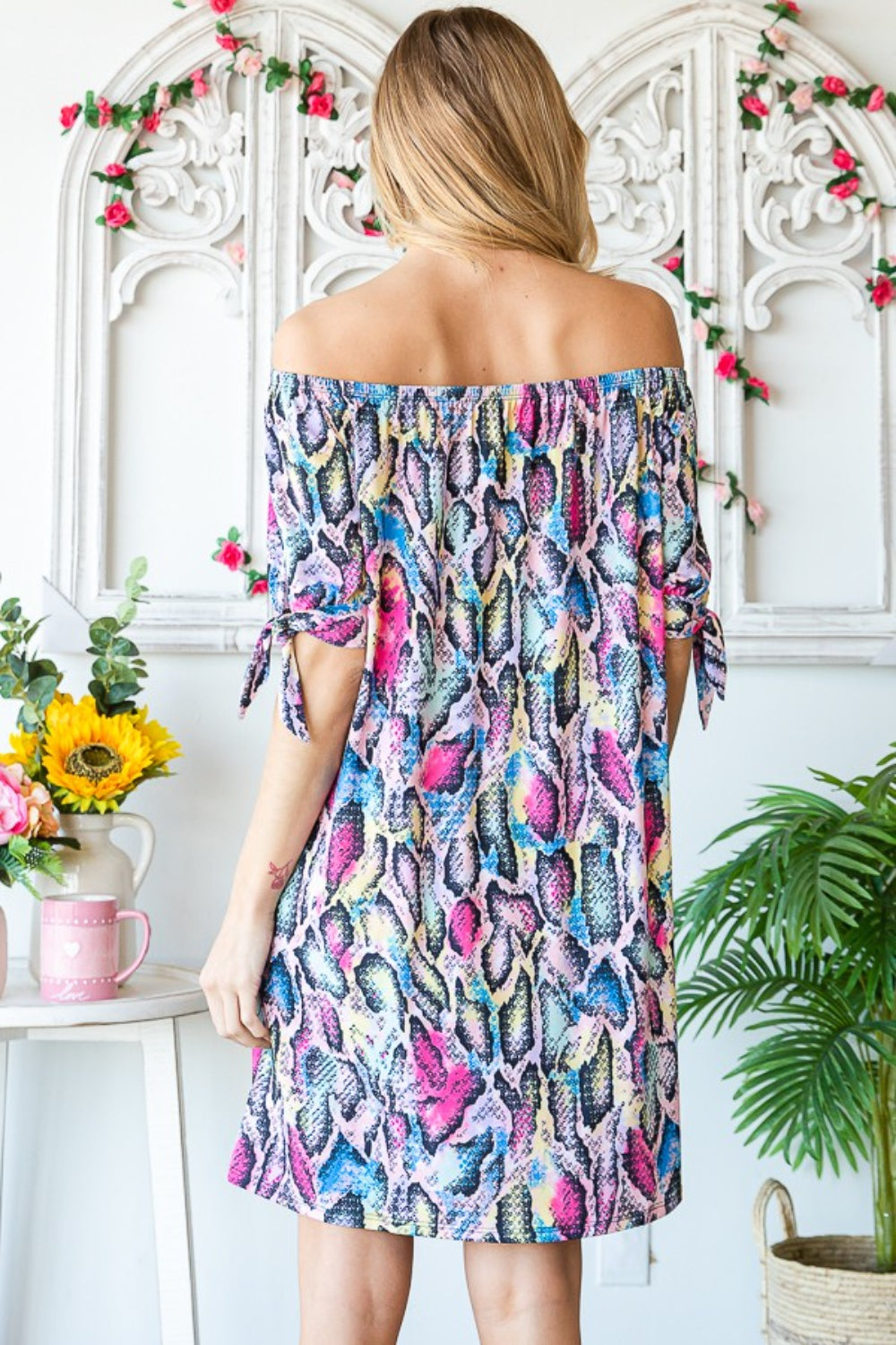 A woman in a stylish Heimish Full Size Off Shoulder Snake Mini Dress and a large straw hat stands next to indoor plants.