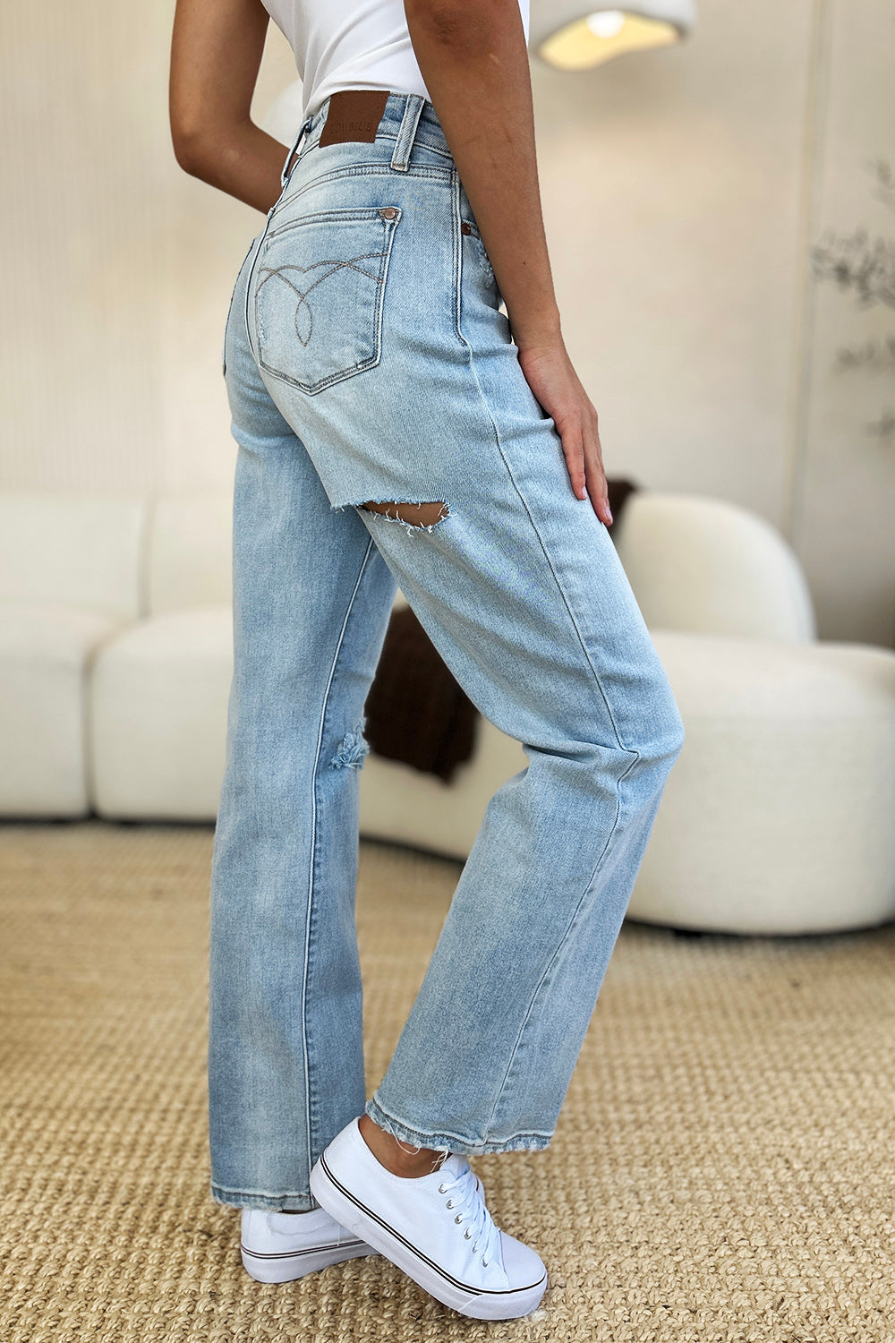 A person standing on a beige carpet, wearing Judy Blue Full Size High Waist Distressed Straight Jeans in light blue, a white top, and white sneakers.