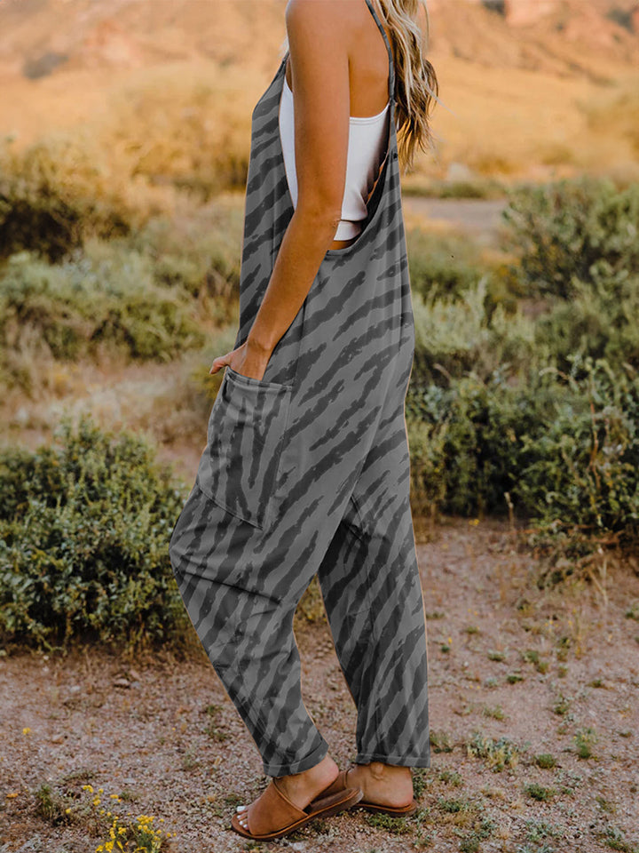 A person wearing a casual white V-neck tank top and a Full Size Printed V-Neck Sleeveless Jumpsuit with a brown animal print zebra-stripe pattern stands outdoors with one hand on their hat and the other in their pocket, set against a rocky landscape backdrop.