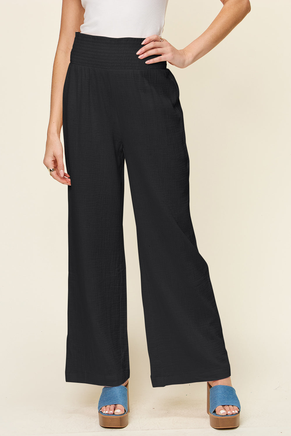 A person wearing Double Take Full Size Texture Smocked Waist Wide Leg Pants in black and a white 100% cotton top is standing with one hand on their hip. They are also wearing blue open-toe sandals.